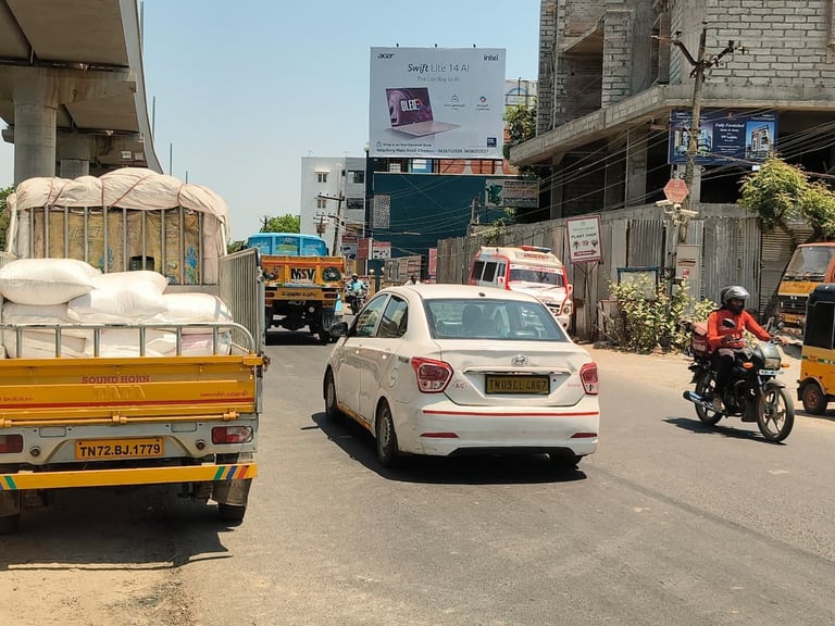 Busy Indian street scene with white car, colorful trucks, and motorcycle amid urban warehouses and industrial buildings