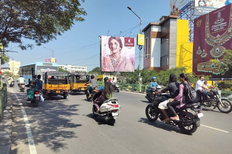Busy urban street with motorcycles, auto-rickshaws, and pedestrians passing modern buildings with large advertisement billboards