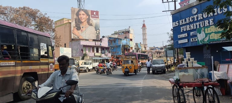Busy Indian street intersection with auto-rickshaws, motorcycles, trucks, and pedestrians under blue sky with buildings and billboards